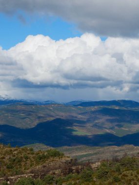 Geniş bir dağ manzarası yeşil tepeler, gölgeli vadiler ve uzak tepelerle açılır. Kabarık bulutlar yukarı doğru sürükleniyor, bu huzurlu doğal ortamın güzelliğini arttırıyor..