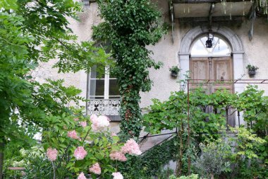 Eski bir evde ahşap bir kapı ve tırmanma bitkileriyle süslenmiş güzel bir balkon bulunur. Bereketli bahçe canlı çiçekler ve yeşilliklerle dolu. Huzurlu bir atmosfer yaratıyor..
