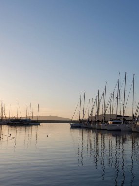 Yat limanına yanaşan yelkenliler gün batımının son ışığını yakalıyorlar. Yansımaları sakin suda parıldıyor. Akşam yaklaşırken, ortalık sakin bir atmosferle çevrili..