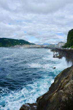 Engebeli bir kıyı şeridi kayalara çarpan dalgaları barındırırken, kıyı kenti bulutlu bir gökyüzünün altında dramatik bir doğal manzara sunuyor..