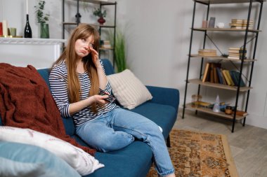 Televizyon izleyen üzgün kadın elinde uzaktan kumandayla koltukta oturuyor.