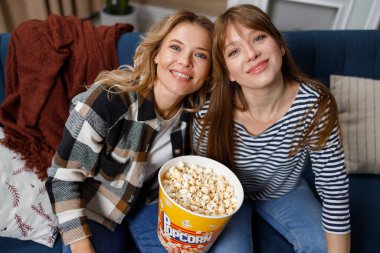 Yetişkin bir anne ile yetişkin bir kızın film izlerken elinde patlamış mısırla kanepede oturmasının portresi.