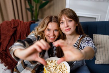 İki mutlu kadın ellerinde patlamış mısırla kanepede otururken kalp işareti yapıyor.
