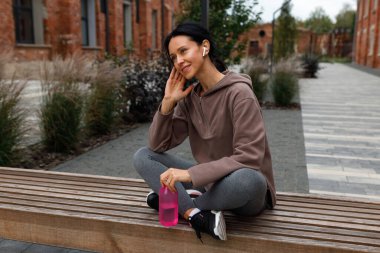 Spor elinde bir şişe suyla güzel bir fitness kadını dışarıda antremandan sonra bankta otururken kulaklıkla motivasyon müziğinin keyfini çıkarıyor.