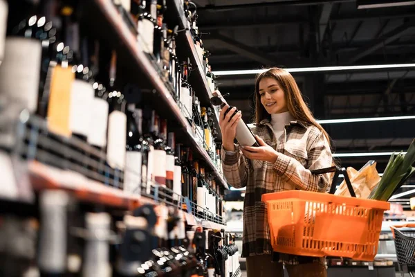 Elinde bir sepet yiyecek olan kadın bir süpermarketin alkol reyonundan şarap seçiyor.