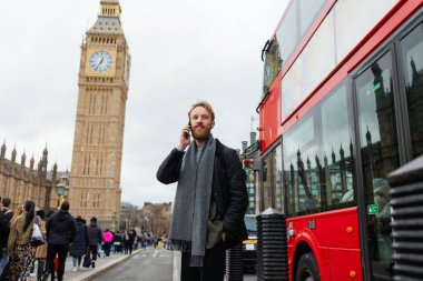 30 'lu yaşlarda sakallı bir adam, Big Ben' in yanında, yoldan geçen kırmızı bir otobüsün yanında konuşuyor.