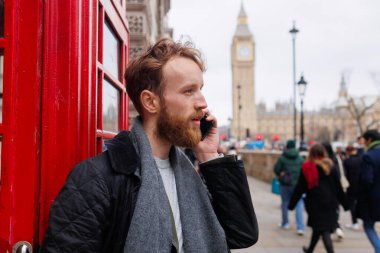 Adam, bir Londra caddesinde kırmızı bir telefon kulübesinin yanında Big Ben kulesinin arka planında dikilirken akıllı bir telefonla konuşuyor.