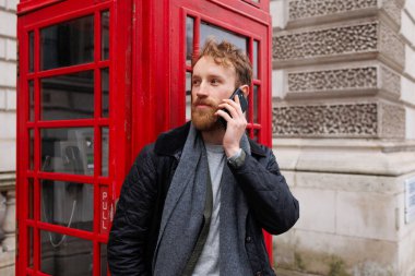 Sakallı adam, kırmızı telefon kulübesinin arka planında akıllı bir telefonla konuşuyor.