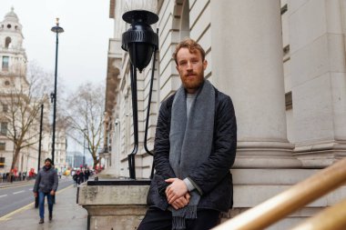 Sakallı, ceketli, atkı takmış şık bir adam Londra sokaklarında eski güzel bir binanın yakınında poz veriyor.