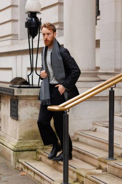 Ceketli ve atkılı şık sakallı adam binadan çıkarken merdivenlerden iniyor.