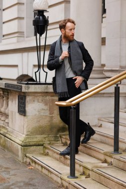 Ceketli ve atkılı şık sakallı adam binadan çıkarken merdivenlerden iniyor.