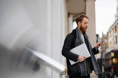 Ceketli ve atkılı sakallı bir iş adamı binanın girişinde dururken akıllı telefon kullanıyor.