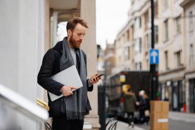 Ceketli ve atkılı sakallı bir iş adamı binanın girişinde dururken akıllı telefon kullanıyor.