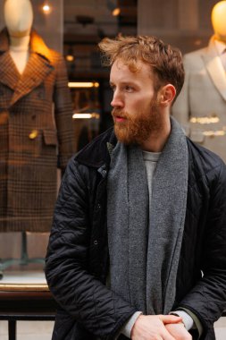 Ceketli ve fularlı sakallı adam elbise mağazasının yanında poz veriyor.