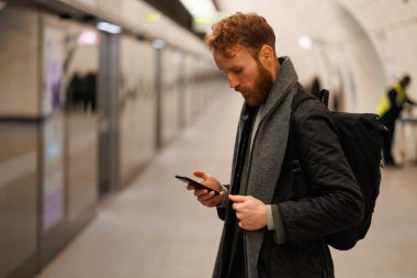 Treni beklerken metro istasyonunda akıllı telefon kullanan bir adam.