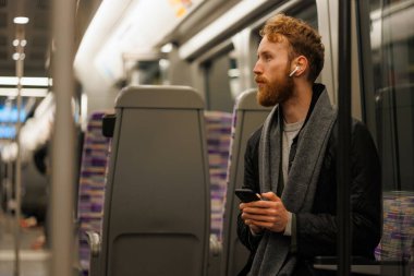 Adam elinde akıllı bir telefonla metroda oturuyor ve kablosuz kulaklık takıyor.