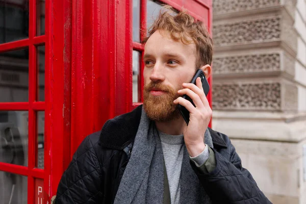 Szakállas Férfi Egy Okostelefonon Beszél Egy Vörös Telefonfülke Hátterében — Stock Fotó