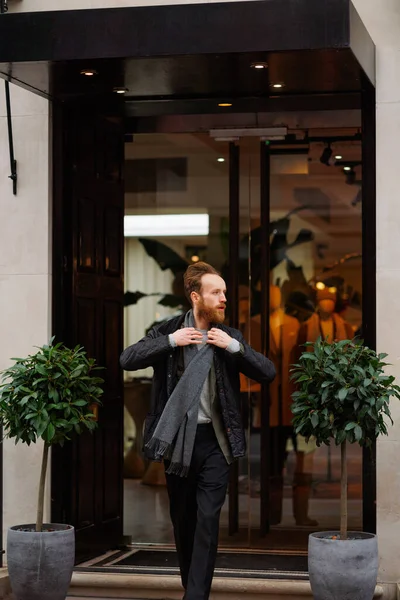 Stylish bearded man in a jacket and scarf goes out of the store or restaurant to the street