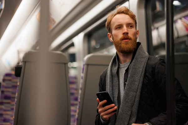 Przystojny Brodaty Mężczyzna Siedzi Pociągu Metra Smartfonem Słuchającym Muzyki Przez — Zdjęcie stockowe
