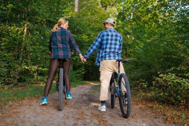 Bisikletli genç çift orman yolunda el ele tutuşuyor.
