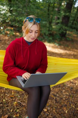 Kırmızı kapüşonlu, kahverengi taytlı ve ormanda hamakta dizüstü bilgisayarı olan hoş, genç, kızıl saçlı bir kadın.