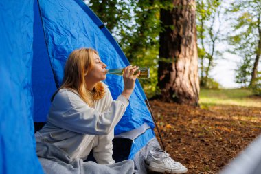 Kız ormanda çadırda otururken bira içer.