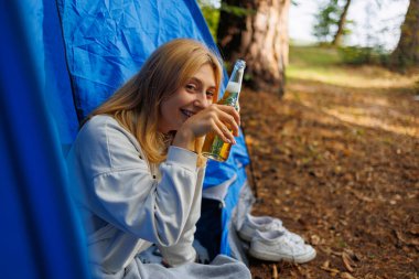 Elinde bir şişe birayla ormandaki çadırda oturan neşeli genç bir kadın.