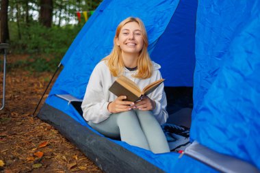 Elinde kitapla gülümseyen mutlu bir kadın ormanla çevrili bir çadırda oturuyor.