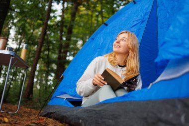 Çadırda elinde kitap olan genç bir kadın ormana bakıyor.