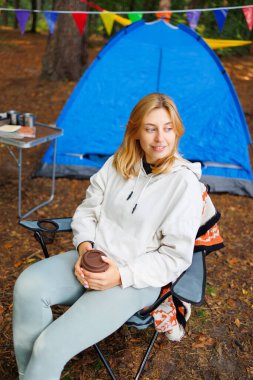 Kapüşonlu ve tozlu bir sandalyede oturan genç bir kadın ormanın ortasındaki bir kamp alanında bir fincan kahveyle arka planda çadır ve karnaval çelenkleri olan.