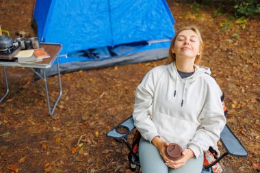 Elinde kahve fincanı olan genç bir kadın kamp sandalyesinde oturur. Çadırın arka planına yaslanır. Gözleri kapalı rahatlar ve açık havanın tadını çıkarır.