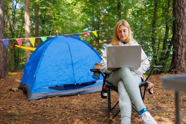 Genç bir kadın ormanın ortasında bir kamp alanında dizüstü bilgisayarla bir sandalyede oturuyor.