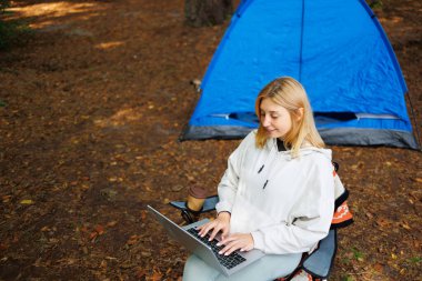 Ormanda kamp yaparken sandalyede oturan ve dizüstü bilgisayarda çalışan bir kadın.