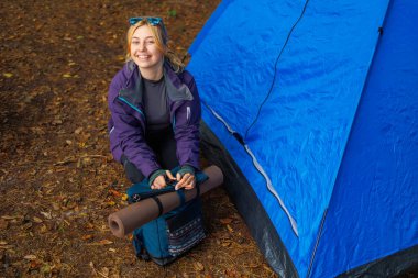 Ormandaki çadırın yanında yürüyüş için hazırlanan mutlu bir bayan yürüyüşçü. Sırt çantasına yoga minderi yerleştirip hazırlanıyor.