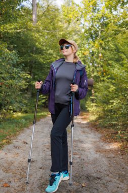 Spor kıyafetli genç bir kadın yürüyüş direğiyle orman yürüyüşünden zevk alıyor.