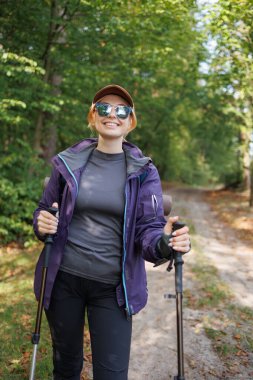 Yürüyüş direkleri ile orman yolunda yürüyen gülümseyen kadın yürüyüşçü.