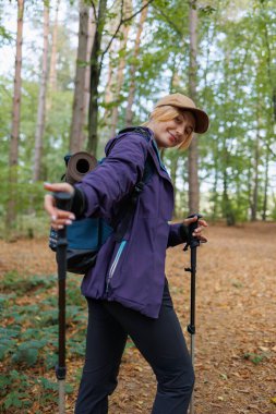 Sonbahar ormanında sırıkla yürüyen ve kameraya gülümseyen bir kadın.