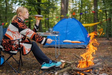 Kamp ateşinde dinlenen, bira içen ve çadırın yanında sosis kızartan bir kadın.
