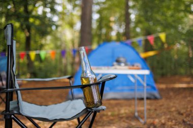 Kamp sandalyesinde bira şişesi ve orman kampında renkli bayraklar.