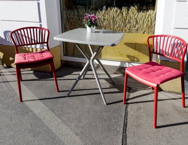 Sabah güneşinde iki kırmızı sandalye ve gri bir masa. Ziyaretçi olmadan.