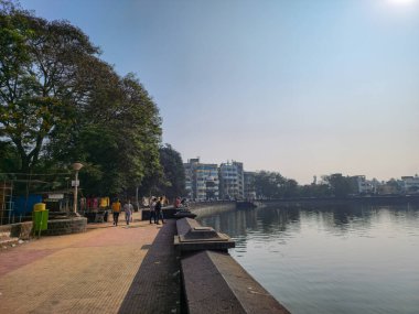 Kolhapur,India- January 27th 2023; Stock photo of ancient beautiful Rankala lake, old stone retaining wall around it. Few people walking, sitting, sightseeing the scenery early in the morning.