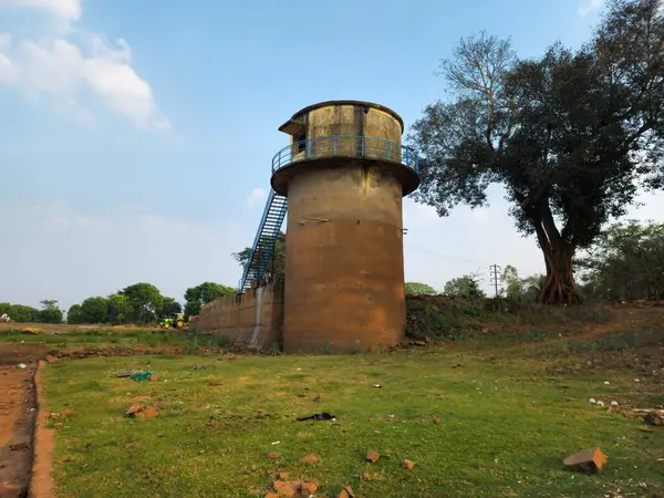 stock image stock photo of A rustic old water tank, connected by iron stairs, surrounded by a landscape covered with green grass , tall trees and a blue sky in the background. Picture captured at kolhapur.