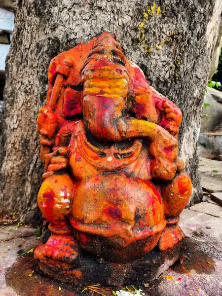 Lord Ganesha 'nın canlı turuncuya boyanmış, eski, rustik bir heykelinin resmi, engebeli bir ağaç kabuğunun arka planına kurulmuştur. Fotoğraf Mailapur, Karnataka, Hindistan 'da çekildi..