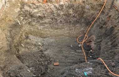 Açık toprak katmanları, turuncu kablo ve inşaat enkazı olan derin bir kazı alanı. Kazılmış kuyu jeolojik dokular, gevşek toprak ve altta tek bir tuğla gösteriyor..