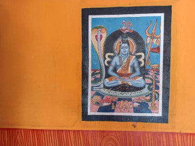 Hindu tanrısı Shiva 'yı tasvir eden çerçeveli bir sanat eseri canlı turuncu bir duvara asılır. Shiva, Trishul (mızrak), bir kobra ve başının üstündeki kutsal Om sembolü eşliğinde meditasyonda oturur..