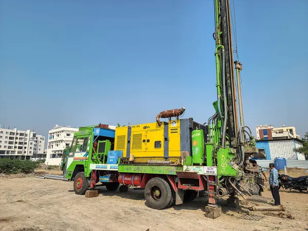 Gulbarga, Hindistan 21 Aralık 2023 Borewell sondaj kamyonu bir inşaat sahasında konuşlandırılmış, uzun yeşil sondaj teçhizatı, sarı Atlas açık mavi gökyüzü altında düzeneği denetleyen bir teknisyen.