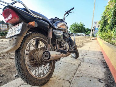 Gulbarga, Hindistan, 20 Ağustos 2023. Tozlu, siyah, görkemli bir motosiklet manzarası açık gökyüzünün altında, ağaçlar, binalar ve arka planda sarı renkli bir duvar bulunan fayanslı bir kaldırımda park edilmiş..
