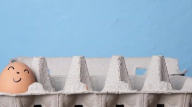 Mavi arka planda yumurta tepsisine gülümseyen yumurtalar katlıyor. Paskalya konsepti. Paskalya 'nın kültürel sembolü. Yüzleri çizilmiş çiftlik yumurtaları..