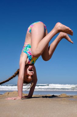 Teen happy and smiling girl performs gymnastic and dance exercises and poses against the backdrop of the sea and beach. The girl stands on her hands clipart