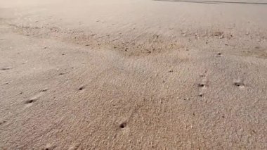 İki çocuk plajda koşuyor. Koşan bacaklara yakın görüş. Kumsaldaki kumda ayak izleri. Kamera kumsaldaki büyük kum tepeciklerindeki genç kız ve kız çocuklarının ayak izlerini takip ediyor.